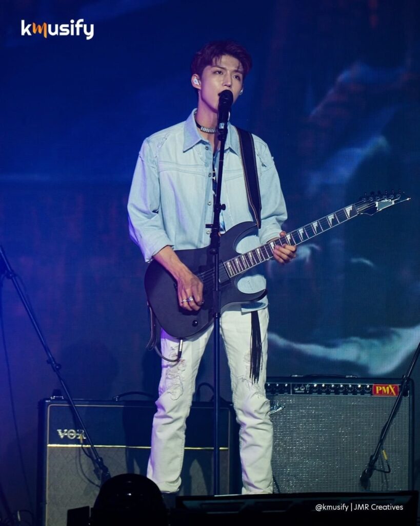 Xdinary Heroes band member Jun Han in white jeans performing on stage, singing and strumming a black guitar.