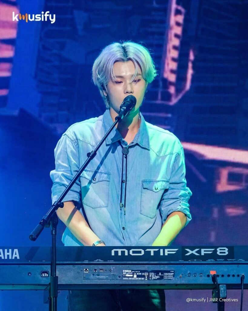 Xdinary Heroes Jung Su wearing a denim shirt, singing while playing the keyboard.