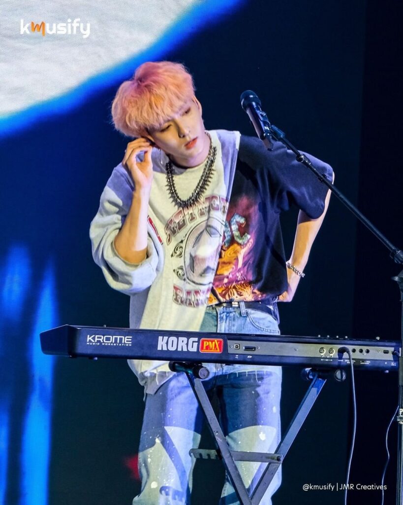Xdinary Heroes band member with pink hair, Od.e, behind an electronic keyboard, removing his ear piece to listen to the crowd's cheers.